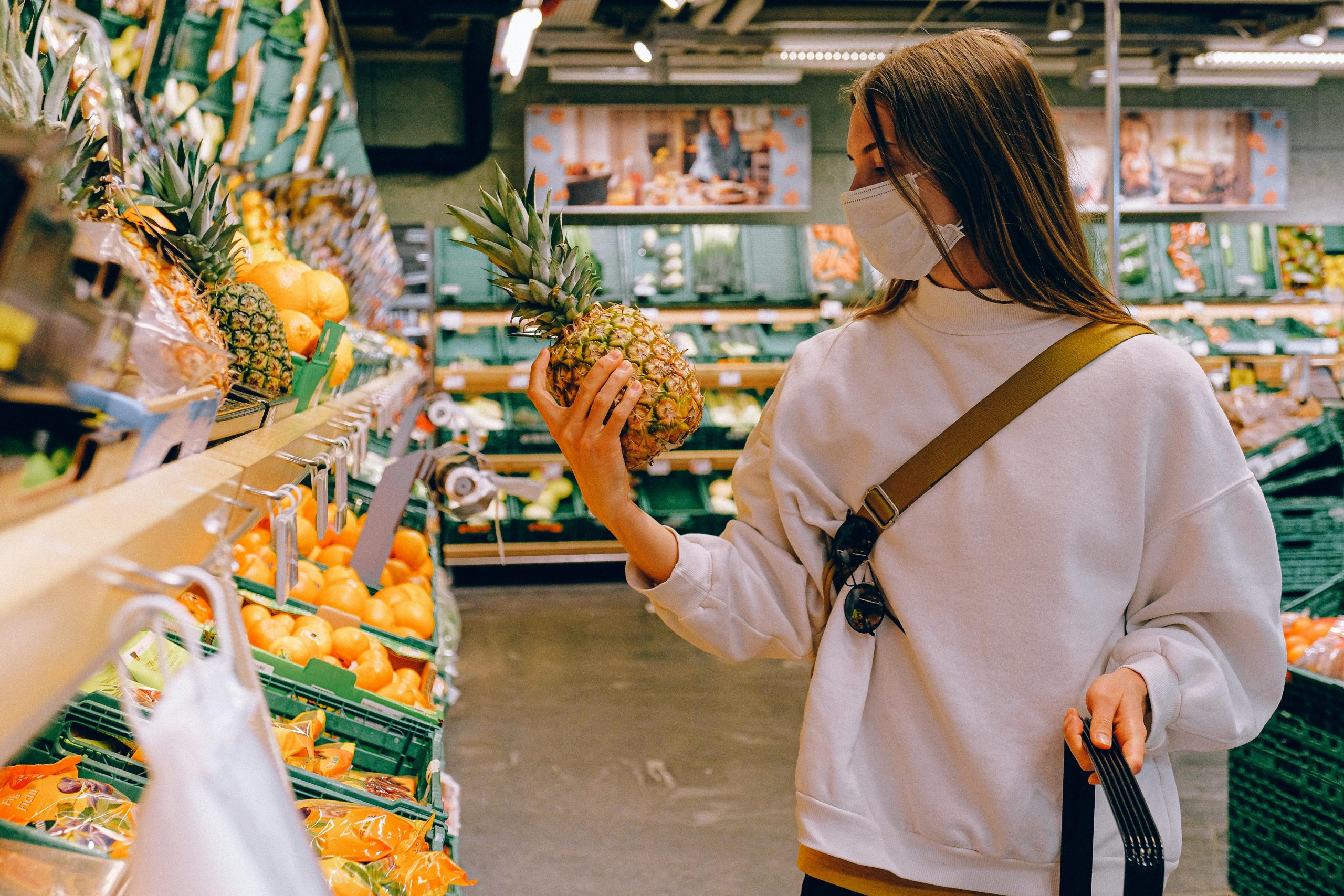 Woman in mask chooses pineapple in a supermarket for safe shopping during pandemic.